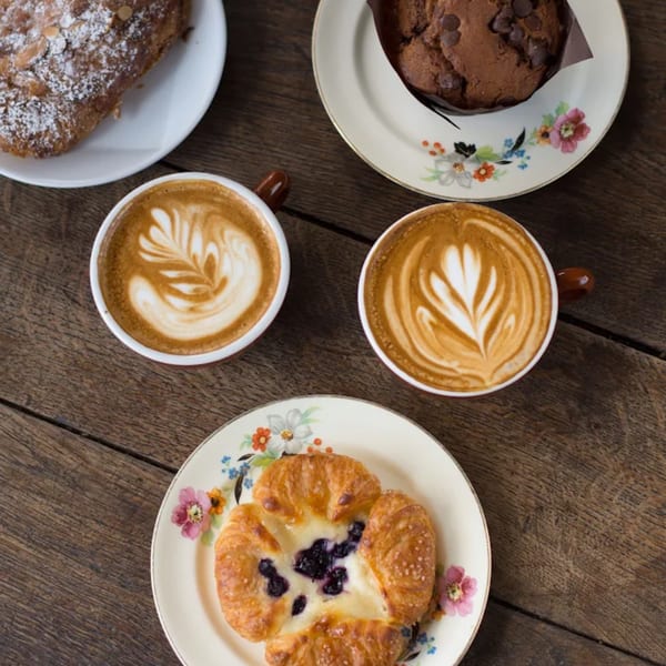 Overhead view of two lattes with latte art, a chocolate chip muffin, and two pastries on a wooden table.