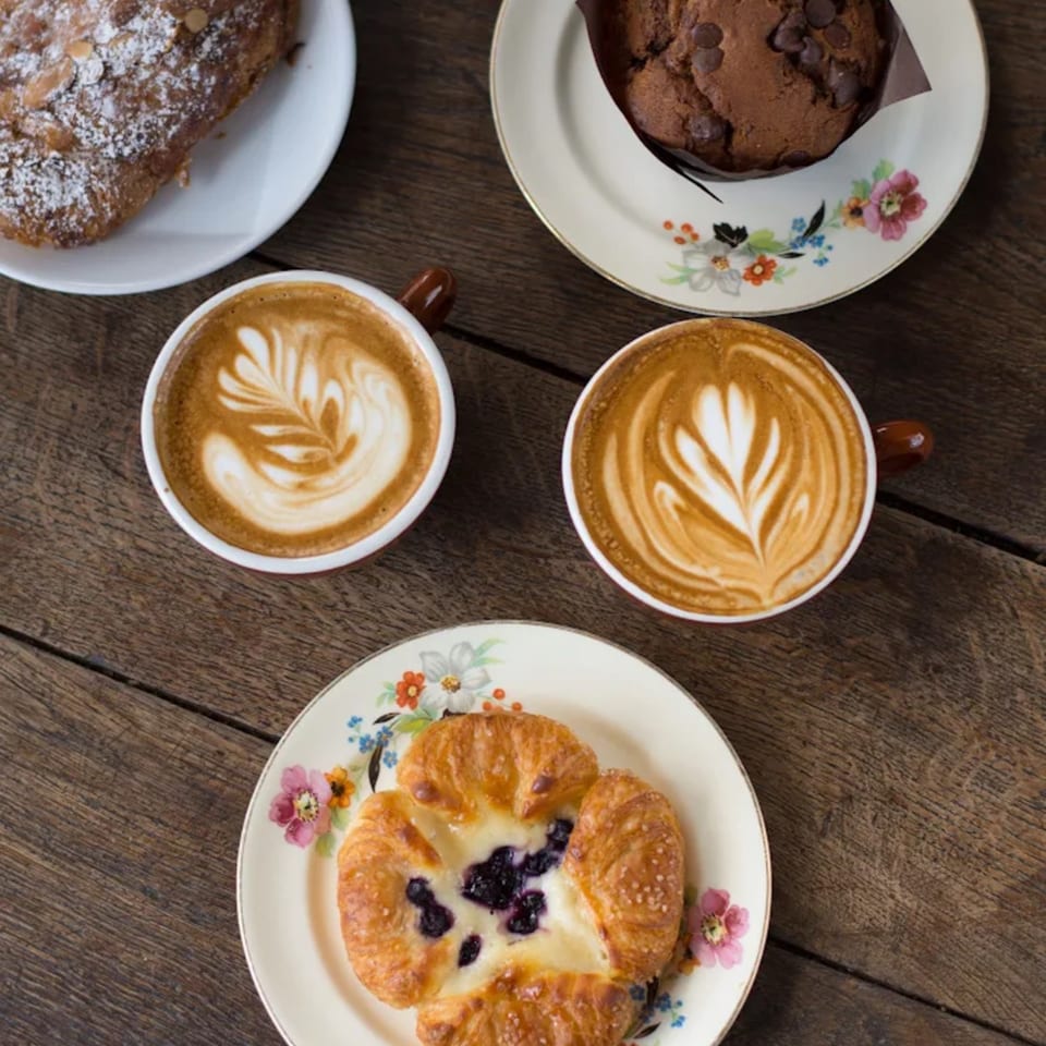 Overhead view of two lattes with latte art, a chocolate chip muffin, and two pastries on a wooden table.