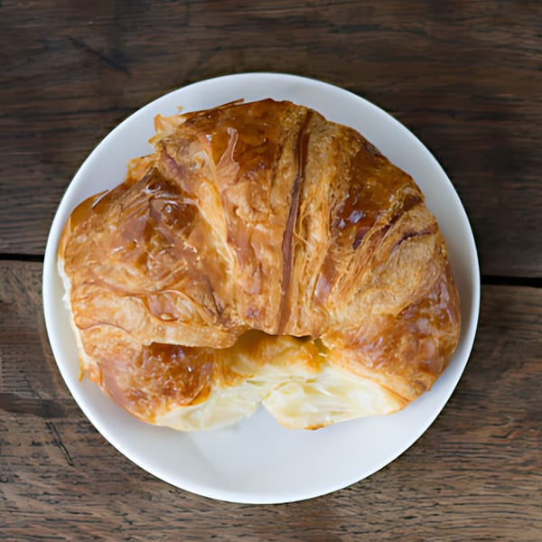 Golden-brown croissant on a white plate, showcasing its flaky layers, against a rustic wooden table.