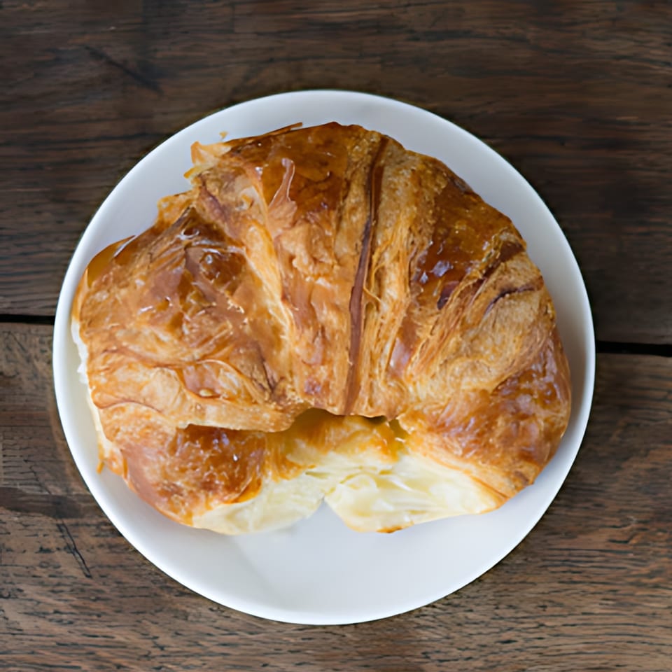 Golden-brown croissant on a white plate, showcasing its flaky layers, against a rustic wooden table.