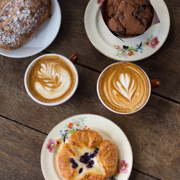 Overhead view of two lattes with latte art, a chocolate chip muffin, a blueberry danish, and a pastry.