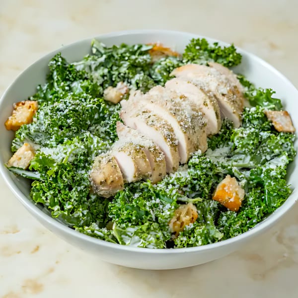 A white bowl filled with a kale Caesar salad, topped with sliced chicken breast and croutons.
