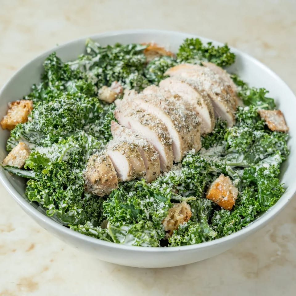 A white bowl filled with a kale Caesar salad, topped with sliced chicken, croutons, and parmesan.