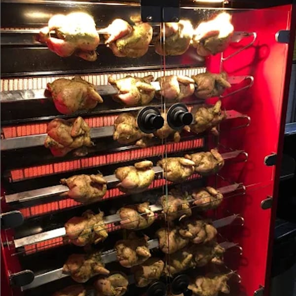 Many whole chickens roast on rotisserie spits inside a red and black oven at Roast & Co.