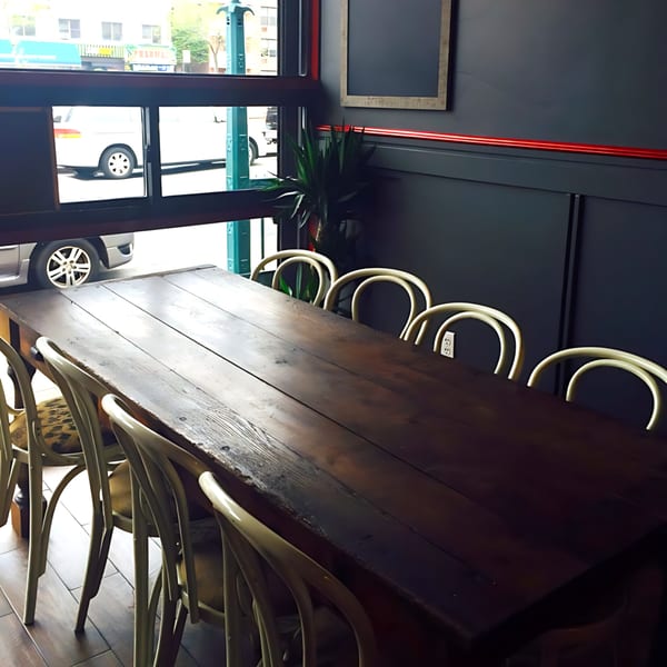 Long wooden table with light-colored chairs in a restaurant dining area with large windows.